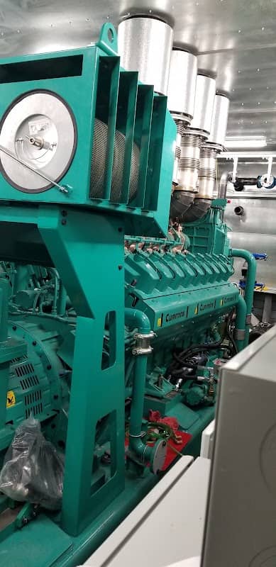 The inside of a large green Cummins gas generator showing the engine block, the exhaust manifold and the pipework above it
