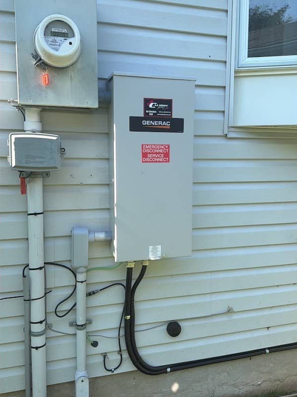 A Generac automatic transfer switch mounted on house siding beside the hydro meter, labelled as the emergency service disconnect, with conduit running down to grade