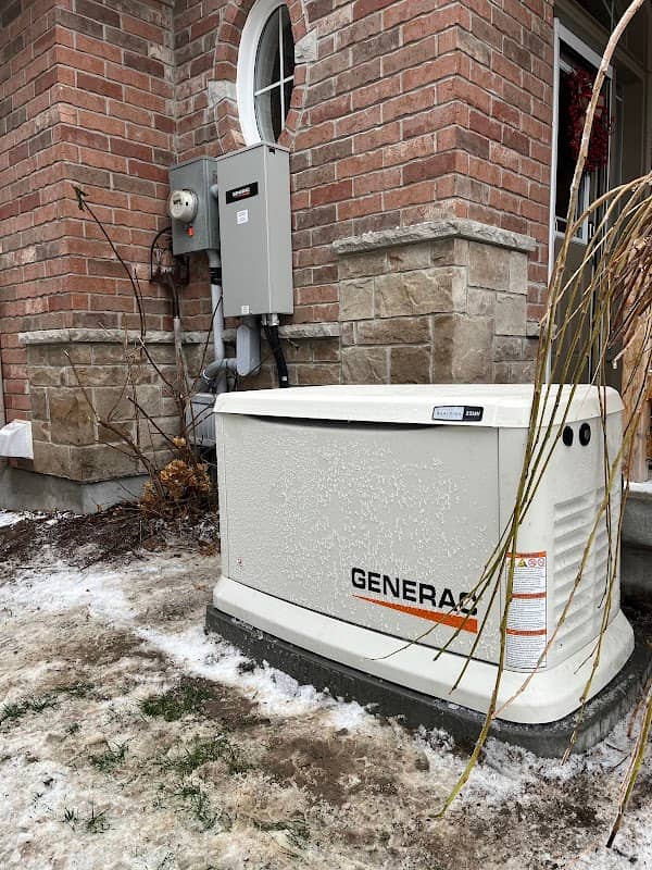 A Generac standby generator on a pad in snow beside a brick house, with the hydro meter and disconnect mounted on the wall above it